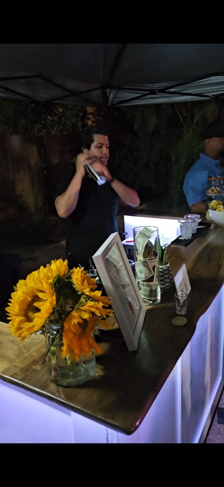 Bartender shaking cocktail shaker at outdoor bar with sunflowers and tip jar in foreground.