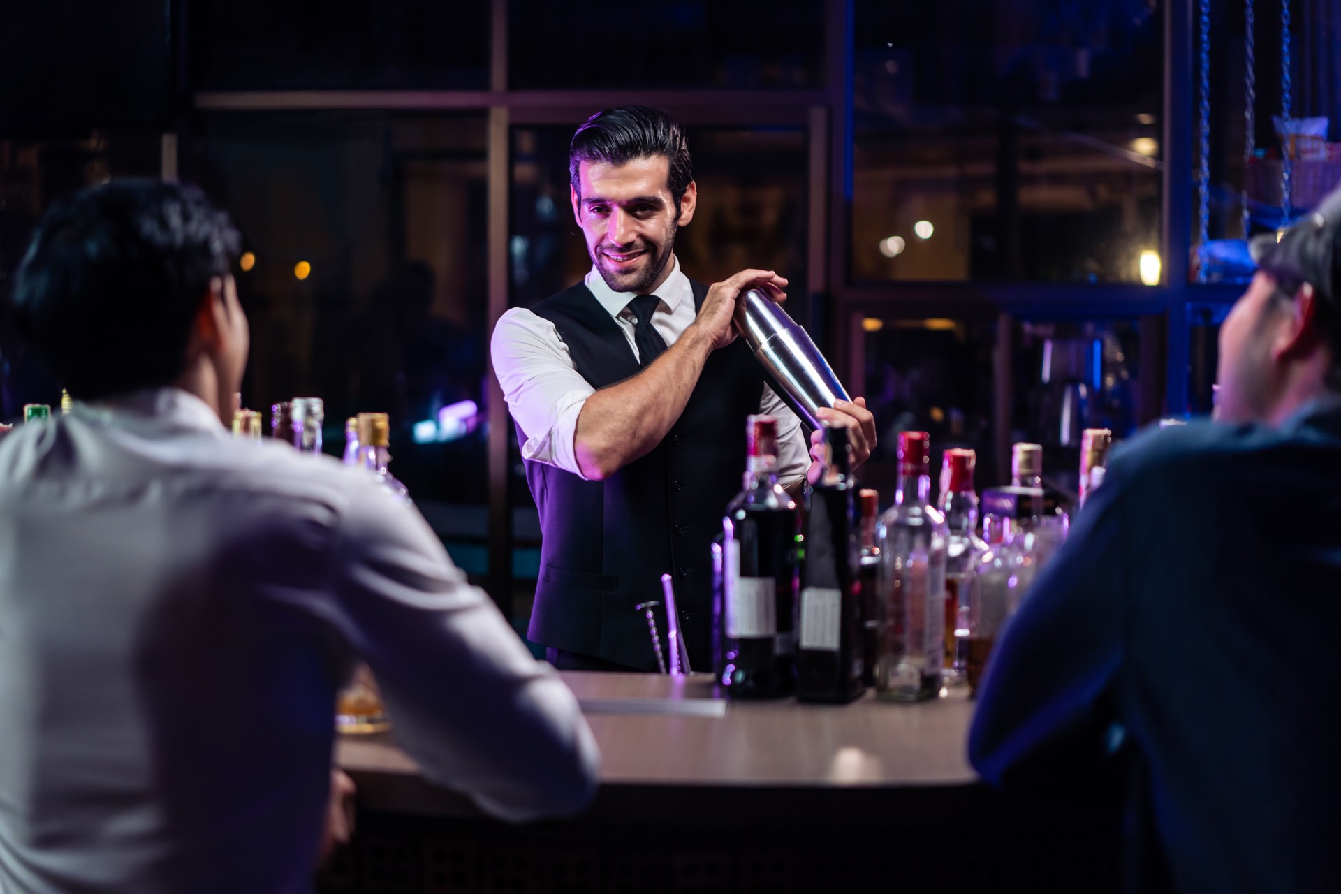 Caucasian profession bartender making a cocktail for women at a bar. Attractive barman pouring mixes liquor ingredients cocktail drink from cocktail shaker into the glass at night club restaurant.